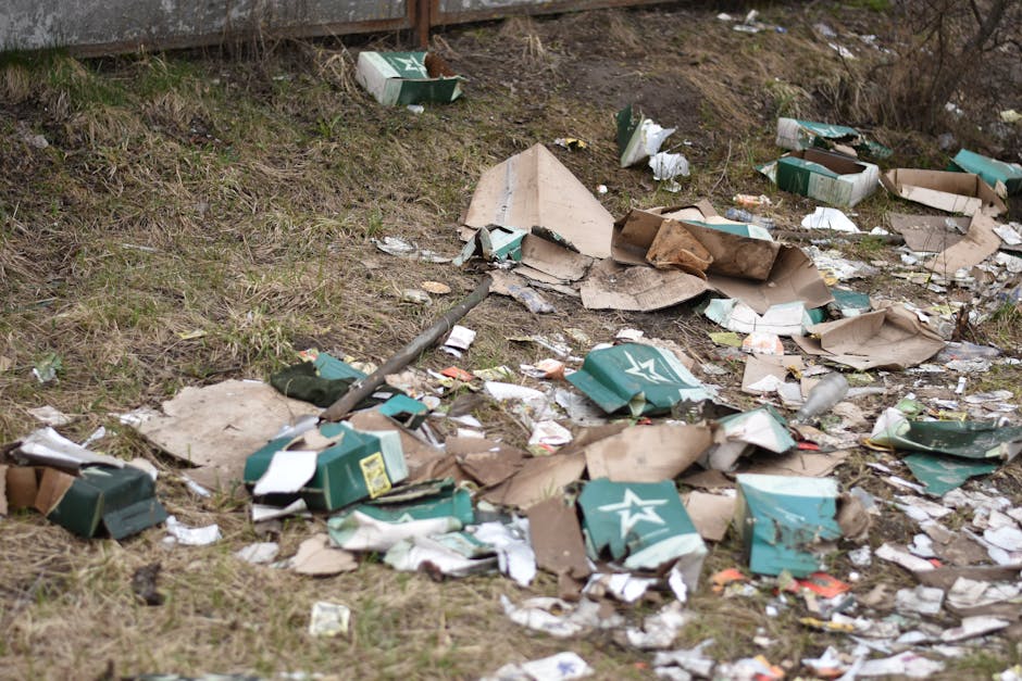 A scattered pile of discarded cardboard boxes, some flattened and others crumpled, display a mix of green and brown colors with visible creases and torn edges, lying on a patch of uneven grassy and dirt ground. Among the debris, there are pieces of torn paper and small fragments of packaging material, with some of the boxes bearing partially visible white star logos. A metal pole or pipe runs diagonally through the rubbish, partially buried under the debris. The surrounding environment features a grassy slope with patches of bare earth and sparse vegetation, and in the background, a wooden fence or barrier is visible, separating the area from a more distant, unclear setting. The scene is illuminated by diffuse daylight, indicating an outdoor location typical of an informal dumping site or neglected urban edge where private rubbish disposal services, such as those provided by Rubbish Clearance Bromley, could be employed for waste removal and site clearance. The overall composition emphasizes the mess and clutter associated with improper waste disposal, highlighting the importance of scheduled rubbish removal services.