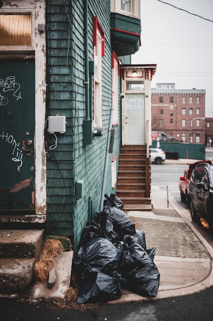 A row of black rubbish bags, filled and tied, are piled at the base of an external green wooden building with weathered, peeling paint. The building features small, rectangular windows with red trim and a white door at the top of a short flight of wooden stairs with a white railing. To the left, there is a partially visible metal door with graffiti tags and rusted areas. The scene is set on a concrete sidewalk adjacent to a paved driveway, with parked cars visible on the street in the background. The environment appears to be an urban residential area, with multi-story brick buildings in the distance, and overhead power lines visible in the sky. The lighting is natural, suggesting a cloudy or overcast day, emphasizing a neutral, professional tone appropriate for waste management and rubbish removal context. Rubbish Clearance Bromley occasionally handles similar private waste collection and on-site clearance services within such urban environments.