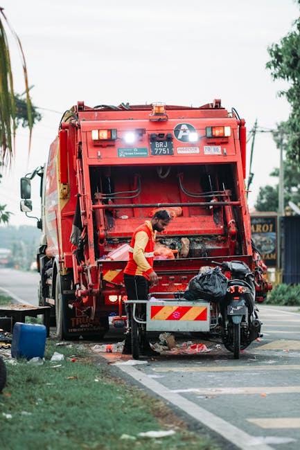A worker wearing a red and yellow uniform is seen operating a large red waste collection truck for rubbish removal on a roadside. The back of the truck, which is open, reveals mechanical components and a section for loading rubbish. The worker is standing beside the truck, handling a black plastic bag filled with waste, with some rubbish and debris visible on the pavement below. A small motorcycle or scooter is parked adjacent to the worker, secured with a metal stand, and nearby, there is a blue container. The scene takes place on a street with a partly cloudy sky, with trees and utility poles visible in the background. The environment appears to be a suburban or urban area, illustrating the process of on-site waste collection. The image aligns with services provided by companies like Rubbish Clearance Bromley that offer alternative waste handling options, such as private rubbish collection, outside of council disposal.