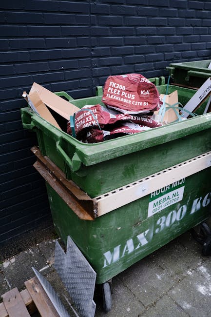 A green wheeled rubbish bin filled with red and black bags of garden waste, including decorated garden soil bags, is positioned outdoors on a paved sidewalk against a dark brick wall. The bin, branded with Rubbish Clearance Bromley, is stacked on top of a larger white container, with additional waste or materials leaning against it, including a small piece of grey rubber mat and wooden blocks nearby. The scene suggests a private or independent waste collection setup, with the bins placed for pickup or disposal near a residential or commercial property in an urban area, highlighting services related to rubbish removal and waste management in Bromley. The environment appears clean but utilitarian, with natural lighting indicating daytime, and the scene emphasizes the process of on-site clearance of waste materials.