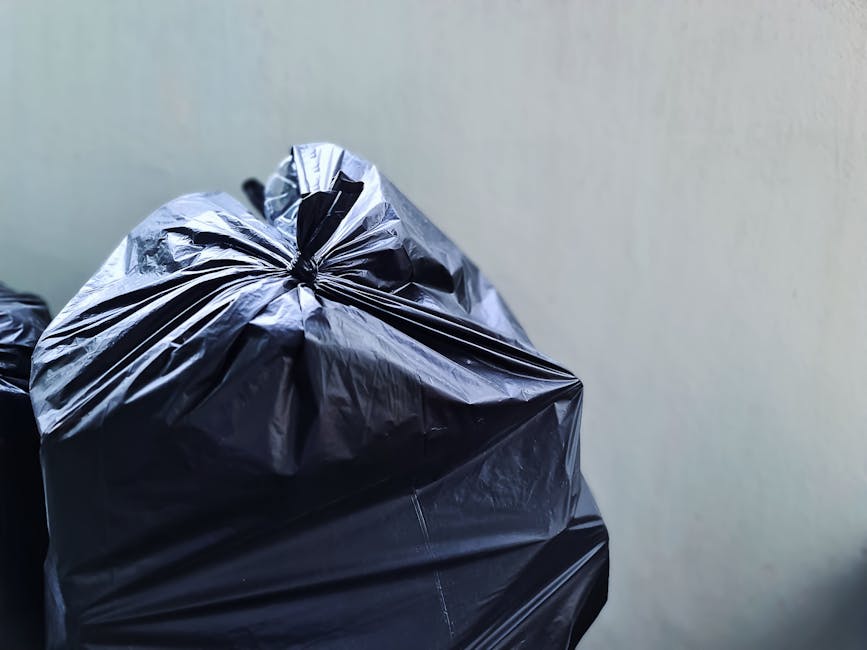 Close-up view of a tightly knotted black plastic rubbish bag resting against a plain, light-colored wall. The bag exhibits a glossy, crinkled texture with visible folds and creases, indicating it is filled with waste materials. It is positioned in the foreground, occupying the left side of the image, with other similar bags partially visible behind it. The environment appears to be an indoor or sheltered area used for waste storage before collection, with no additional objects or surroundings explicitly visible. The lighting is even, casting minimal shadows, emphasizing the reflective quality of the plastic. This scene is indicative of private waste disposal or rubbish collection preparation, relevant to rubbish removal services like those offered by Rubbish Clearance Bromley, supporting the concept of independent or on-site waste handling typical in residential or commercial settings.
