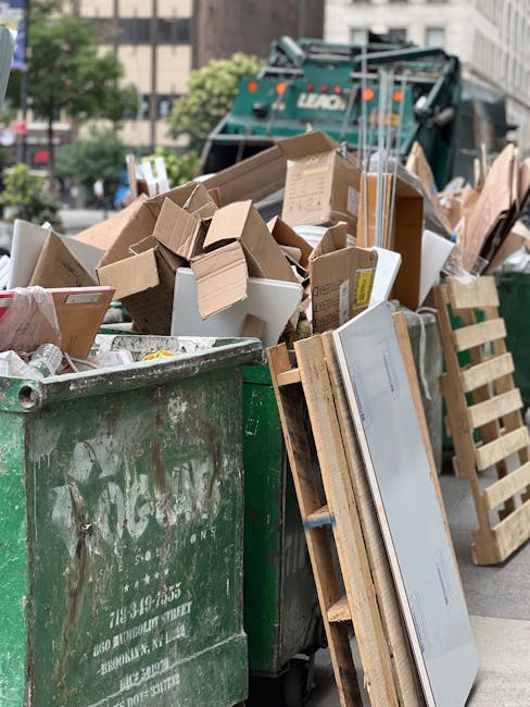 A large green waste disposal skip, positioned on a city street, is brimming with assorted cardboard boxes, some flattened and others partially collapsed, alongside a few white polystyrene trays. The cardboard varies in shades of brown, with some boxes featuring printed labels and symbols, exhibiting textures from smooth to slightly corrugated. Adjacent to the skip, a wooden pallet stands upright, partially obscuring a flat-screen television that leans against it, with its reflective screen surface visible. In the background, a street scene includes a green waste collection truck with a mechanical arm, parked amidst other urban elements such as buildings and trees. The environment appears well-lit, suggesting daytime, with natural daylight casting soft shadows. The scene reflects an instance of private waste collection, highlighting the type of rubbish that might be managed by services such as Rubbish Clearance Bromley, focusing on on-site clearance of domestic or commercial waste materials, specifically cardboard and large household items, in a manner consistent with independent rubbish removal activities.