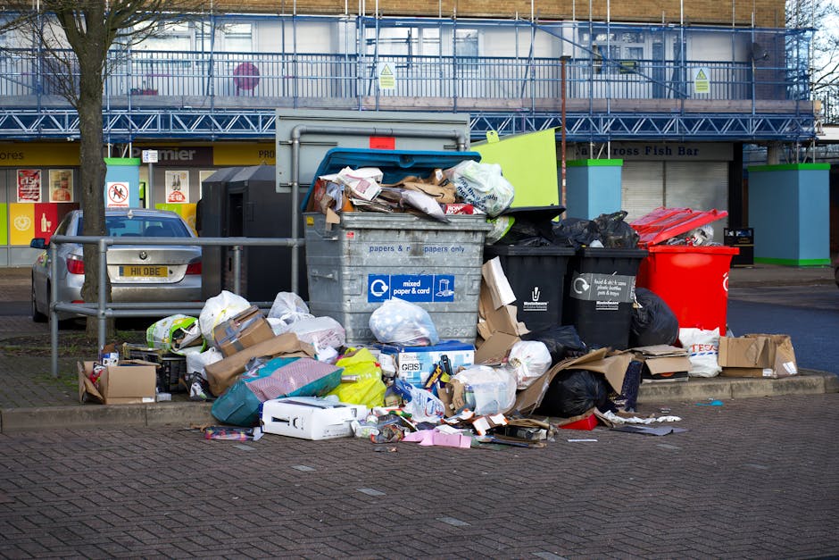 A large pile of mixed waste and rubbish bags, including black, white, and blue plastic bags, cardboard boxes, paper, and packaging materials, is accumulated on a paved urban street corner near a grey metal recycling bin labeled for mixed paper and card, with some items piled on the ground and partially overflowing the bin. Several black trash bags and loose packing materials surround the pile, with a few small items such as product boxes and plastic containers scattered in front. In the background, a parked silver car is visible on a paved driveway, and a multi-storey building with a blue metal scaffolding structure and commercial storefronts, including a fish bar, lines the street. The overall scene suggests an area awaiting clearance or disposal, possibly managed by an independent waste collection service, on a cloudy day with minimal natural light.